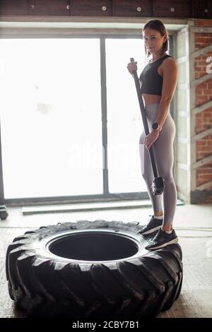 Femme Fitness vêtus de noir et argent haut leggins posant avec une masse, debout près de gros camion pneu dans la salle de sport. Banque D'Images