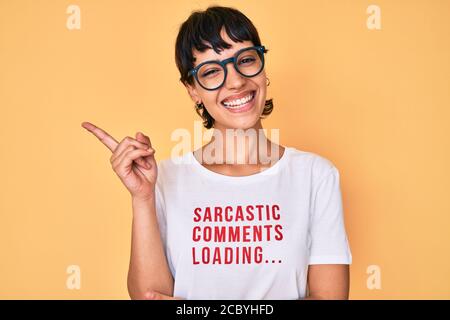 Belle femme brunettte portant des commentaires sarcastiques chargement du t-shirt sourire heureux en pointant avec la main et le doigt sur le côté Banque D'Images