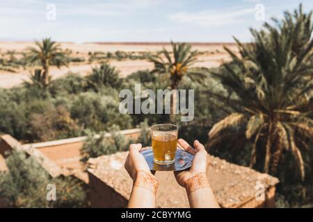Femme mains avec tatouage au henné tenant un verre de thé marocain à la menthe traditionnel. Sable désert et palmiers sur fond. Banque D'Images