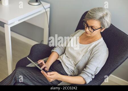 Communication en ligne sur l'éducation au travail. La femme âgée avec des lunettes utilise une tablette à la maison. Banque D'Images