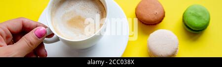 Main de femme tient une tasse de café, macaron de gâteau sur table jaune d'en haut. Bureau de travail pour femme. Petit déjeuner confortable.une tasse blanche de café savoureux dans une femme Banque D'Images