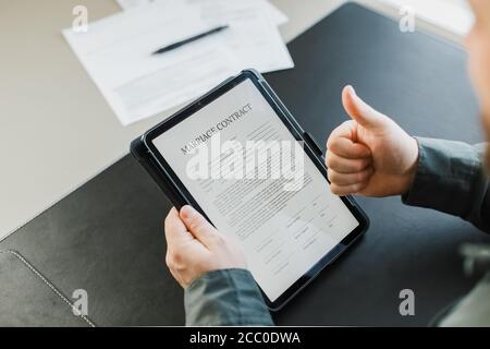 Homme lisant le contrat de mariage sur une tablette au bureau et montrant les pouces vers le haut. Banque D'Images
