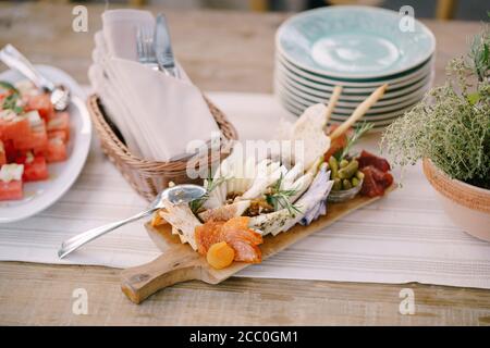 Planche en bois pour couper des produits avec du fromage, des saucisses et des cornichons sur la table avec des assiettes propres, un set de couverts et une pastèque en tranches. Banque D'Images
