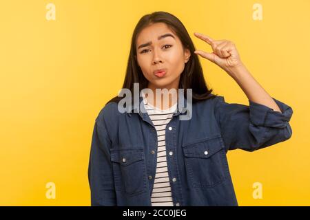 Trop petit ! Portrait d'une fille mécontente dans une chemise en denim montrant un petit geste, insatisfait avec la taille de mesure basse échelle, le pouce minimum ou centim Banque D'Images