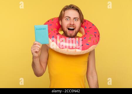Joyeux optimiste drôle de touriste gars dans undershirt debout avec anneau en caoutchouc donut rose autour de la tête et tenant le passeport document, visa gratuit de voyage abro Banque D'Images