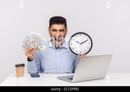 Le temps, c'est de l'argent ! Un homme d'affaires agressif assis sur le lieu de travail de bureau, tenant une grande horloge et des billets de banque en dollars, criant avec colère, en apparence folle furieuse. Banque D'Images