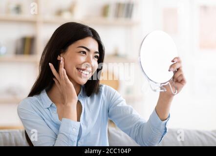 Beauté naturelle. Fille asiatique tenant un miroir grossissant et touchant sa peau parfaite Banque D'Images