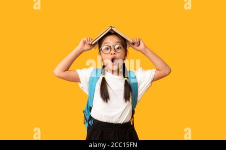 Drôle de fille d'école posant avec livre sur la tête, fond jaune Banque D'Images