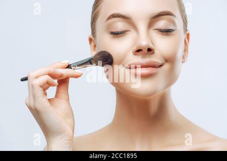 Gros plan portrait de belle fille a la peau fraîche et saine, ferme les yeux, applique des cosmétiques sur le visage, utilise la brosse de beauté, a le maquillage professionnel, pose Banque D'Images