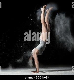 jeune femme à la figure athlétique vêtue d'un body noir se dresse à l'envers sur ses mains, fond noir avec de la farine blanche volante, acrobatique e Banque D'Images