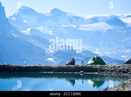 Vue latérale du voyageur de sexe masculin assis près d'une tente touristique près du magnifique lac bleu. Homme parlant à la radio et appréciant la vue sur les montagnes majestueuses. Concept de voyage, de randonnée et de camping. Banque D'Images