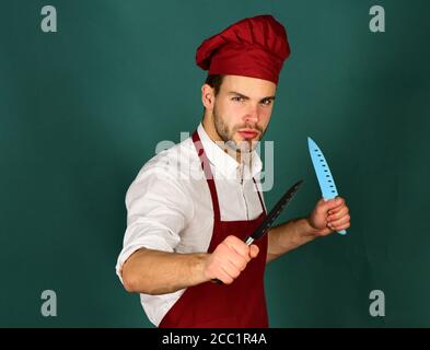 Ustensiles de cuisine et concept de cuisine. Cuisine. Chef au visage sérieux tient des couteaux bleus et métalliques sur fond vert foncé. Homme dans le chapeau de cuisinier et le tablier fouets les couteaux Banque D'Images