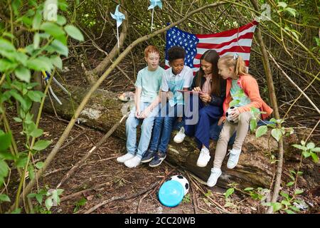 Groupe multiethnique d'enfants utilisant une tablette numérique tout en jouant dans la cour se cachant sous les buissons avec le drapeau américain en arrière-plan, espace de copie Banque D'Images