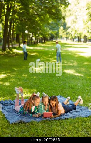 Portrait vertical complet de trois adolescentes utilisant une tablette numérique tout en étant allongé sur de l'herbe verte dans le parc à l'extérieur éclairé par la lumière du soleil, espace de copie Banque D'Images