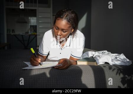 Femme noire faisant des marques sur le document en étant couché sur le lit et étudier à la maison Banque D'Images