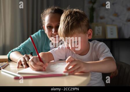Bonne femme regardant un garçon focalisé pour dessiner une image sur la table à la maison Banque D'Images