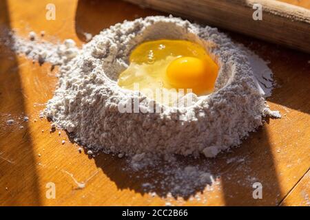 Ingrédients pour faire la pâte. Farine tamisée et œuf cassé sur une table en bois et une broche à roulettes. Photo de haute qualité Banque D'Images