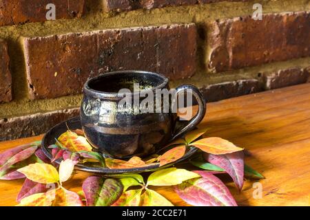 Une composition de vie fixe composée d'une tasse de poterie émaillée et des feuilles d'automne colorées sur un plan d'examen en bois avec un toile de fond en brique Banque D'Images