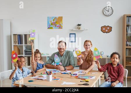 Portrait d'un enseignant souriant avec un groupe multi-ethnique d'enfants montrant des photos de fusées spatiales tout en profitant de l'art et de l'artisanat leçon en préscolaire ou Banque D'Images