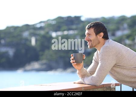 Vue latérale d'un homme heureux contemplant une vue avec un café tasse sur un balcon sur la plage Banque D'Images