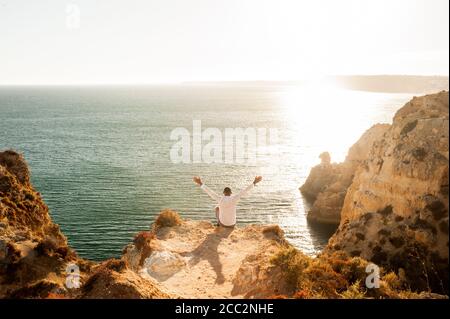 Homme assis sur les rochers avec les bras ouverts au Vue sur l'océan - lumière du coucher du soleil Banque D'Images