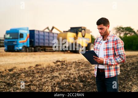 L'agriculteur commande le chargement du blé de la récolteuse au camion à grains. Le conducteur tient le presse-papiers, garde les notes, compte de la cargaison. Le transitaire remplit l'envoi Banque D'Images