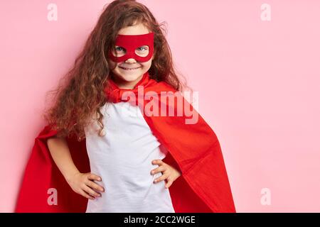 Portrait de super-héros d'enfant souriant confiant portant un manteau rouge et un masque sur les yeux se imagine comme un héros, se tenir isolé ove arrière-plan rose Banque D'Images