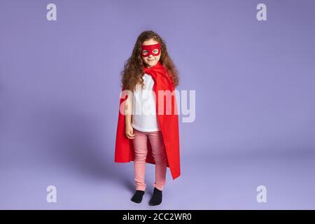 Portrait de petite fille vêtue de cape rouge et de masque, debout isolé sur fond violet Banque D'Images