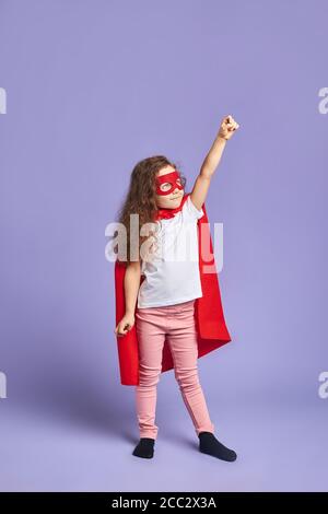 Portrait de petite fille vêtue de cape rouge et de masque, debout isolé sur fond violet Banque D'Images