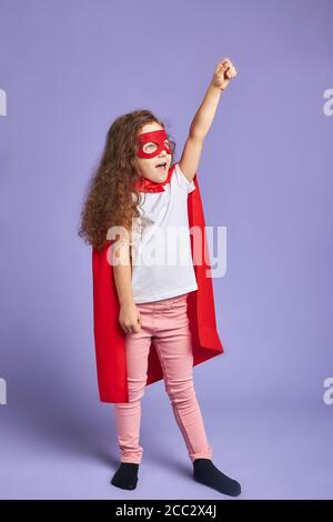 Une petite fille positive a soulevé les bras avec enthousiasme, vêtue de masque rouge et de manteau rouge. Concept Spaceman Banque D'Images