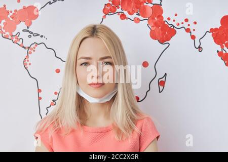 Portrait en gros plan de la triste femme caucasienne dans un masque de protection médicale pendant l'épidémie de Covid-19 dans le monde entier. Carte du monde avec des marques rouges de Coronav Banque D'Images