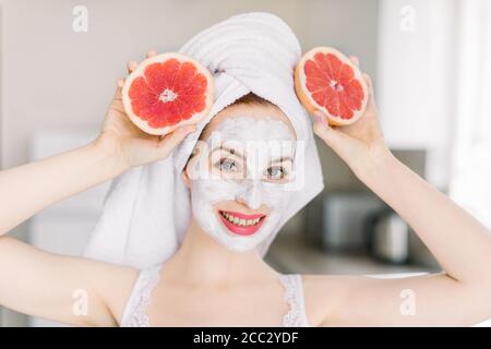 Joyeux sourire jeune fille après la douche, avec masque en argile facial, posant à l'appareil photo sur le fond intérieur de la maison avec des moitiés de pamplemousse frais. Beauté Banque D'Images