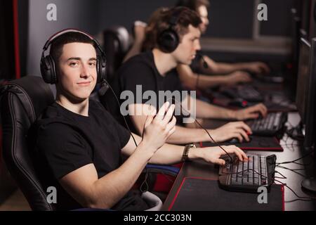 Joueur intelligent satisfait dans un t-shirt noir assis derrière l'ordinateur surveiller et regarder la caméra dans un club de jeu ou internet café avec l'autre peop Banque D'Images