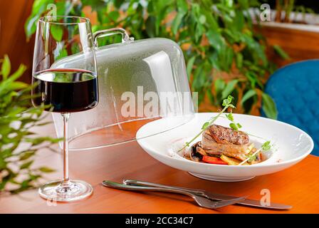 Steak de veau fraîchement cuit avec des légumes et des micro-légumes servis dans une assiette blanche avec un chapeau et un verre de vin. Banque D'Images