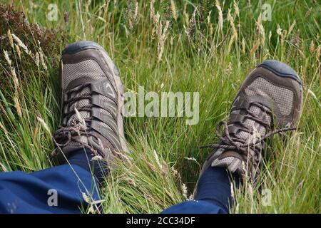 Vue sur les chevilles et les pieds d'un homme, reposant, allongé dans l'herbe rugueuse, portant un pantalon bleu, des chaussettes bleues et des chaussures de marche Banque D'Images