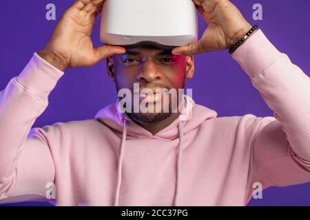 Bel homme africain habillé en blanc à capuche sport, prise casque VR hors de sa tête, d'être impressionné par les effets virtuels, souriant heureusement plus de vi Banque D'Images