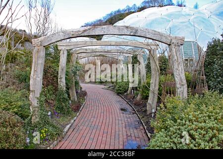 Une allée pavée en brique mène sous quelques anciennes arches en bois sur le chemin d'un biome à l'Eden Project à Cornwall, en Angleterre Banque D'Images