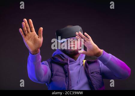 Boxeur professionnel africain en tenue violette décontractée regardant à travers la compétition avec sa participation, en utilisant des lunettes VR, étudiant des erreurs et des cactus Banque D'Images