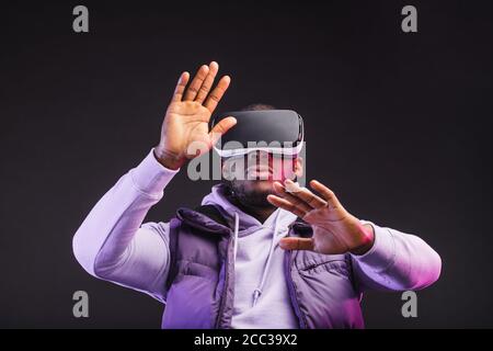 Jeune homme à la peau sombre habillé de vêtements d'extérieur ayant sur la tête Casque de réalité virtuelle mobile pour jouer à des jeux en 3D mais à l'humanité rencontre une nouvelle ère de contro Banque D'Images