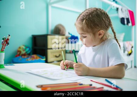 Portrait d'une petite fille mignonne regardant l'appareil photo et souriant tout en dessinant des photos ou en faisant des devoirs, assis à une table à l'intérieur de la maison