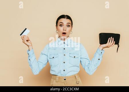 femme brune choquée dans une chemise en denim tenant un portefeuille et un crédit carte isolée sur beige Banque D'Images