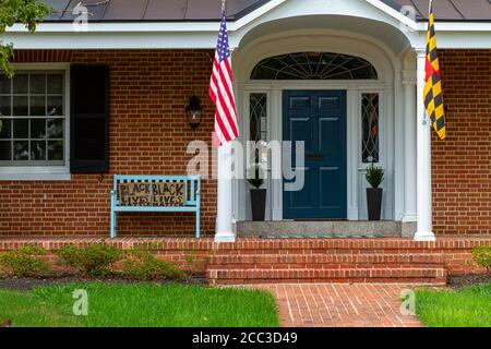 Frederick, MD 08/14/2020: Une maison traditionnelle en brique avec un drapeau américain et un drapeau du Maryland sur les poteaux de drapeau à l'entrée a un banc bleu vintage sur le Banque D'Images