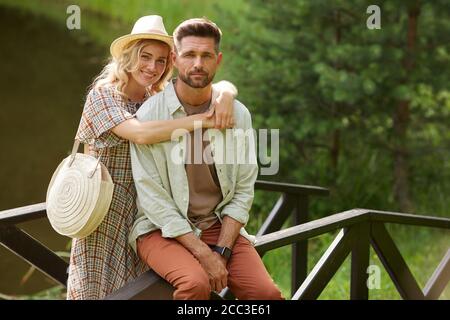 Portrait d'un couple adulte romantique embrassant et regardant l'appareil photo tout en posant sur un pont en bois au bord du lac dans un paysage rustique de campagne, espace de copie Banque D'Images