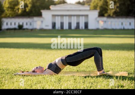 Belle jeune femme couché sur un matelas jaune, posez tout en portant une tenue de sport serrée dans le parc faisant pilates ou yoga, pont d'épaule débutant Banque D'Images