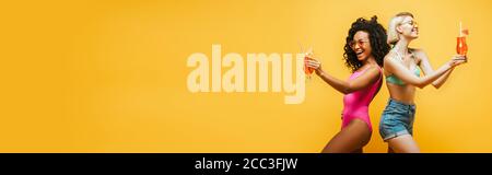 image horizontale de jeunes femmes multiculturelles en tenue d'été debout dos à dos avec des verres à cocktail isolés sur le jaune Banque D'Images