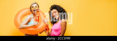 image horizontale de jeunes femmes interraciales avec anneau de natation et verres à cocktail posé isolés sur le jaune Banque D'Images