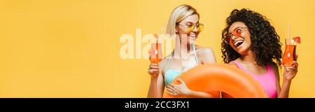 jeunes femmes interraciales en tenue d'été tenant un anneau de natation et verres à cocktail tout en regardant les uns les autres isolés sur le jaune Banque D'Images