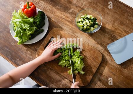 vue de dessus d'une femme coupant de la laitue verte sur la planche à découper près des balances de cuisine et des légumes entiers Banque D'Images