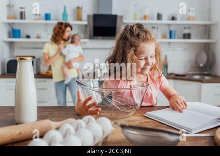 foyer sélectif fille pointant avec le doigt vers le livre de cuisine près du bol, fouet, bouteille de lait et œufs de poulet Banque D'Images