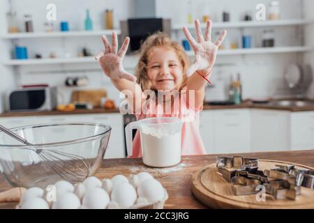 foyer sélectif de la blonde fille montrant les mains dans la farine près de la table de cuisine avec de la farine, des oeufs, des emporte-pièces et fouetter dans un bol Banque D'Images